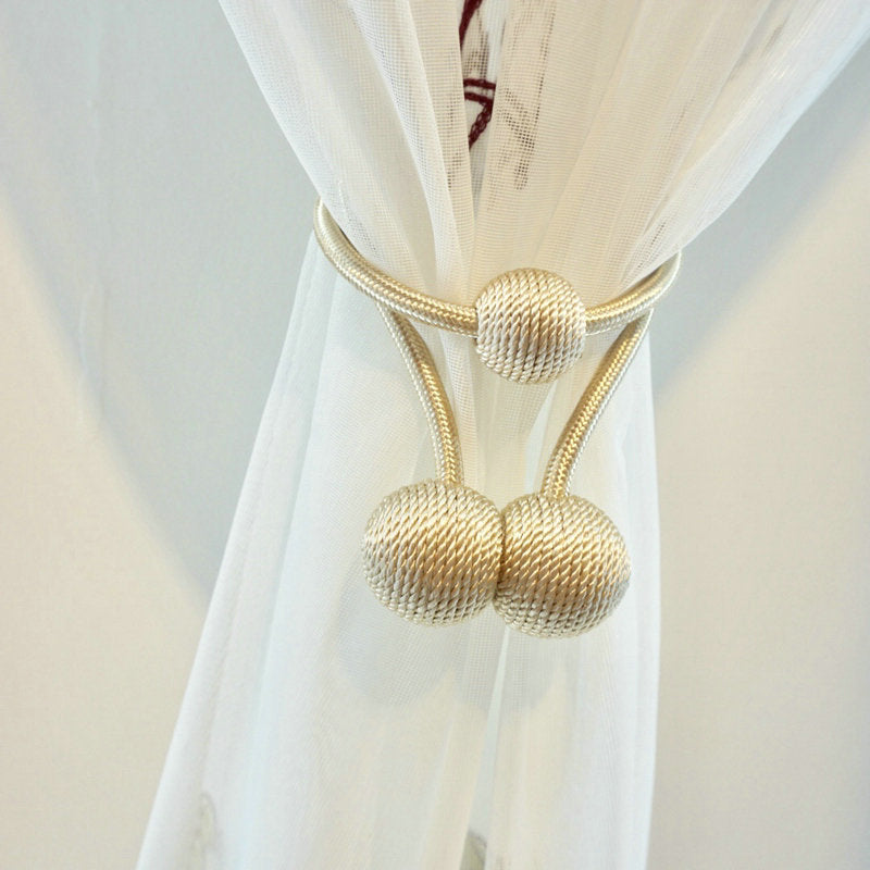 Simple modern gold braided magnetic curtain strap tieback securing a sheer white curtain. Features round woven textured spheres.