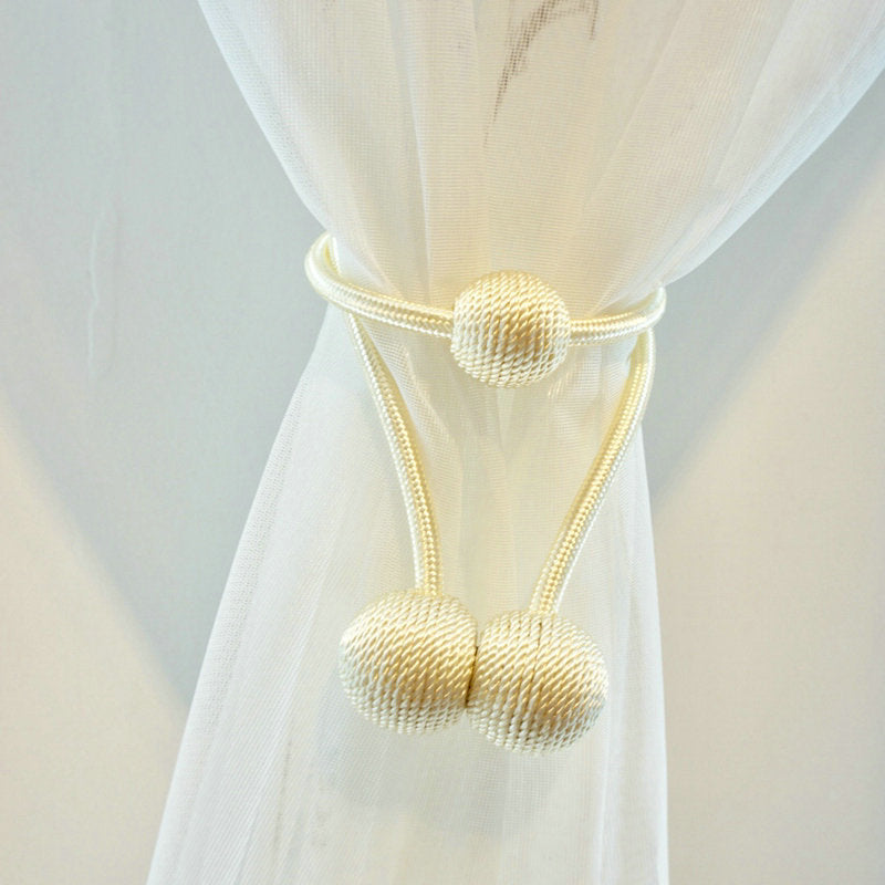 Elegant cream magnetic curtain tieback securing a sheer white curtain. Modern braided rope design with decorative sphere accents.