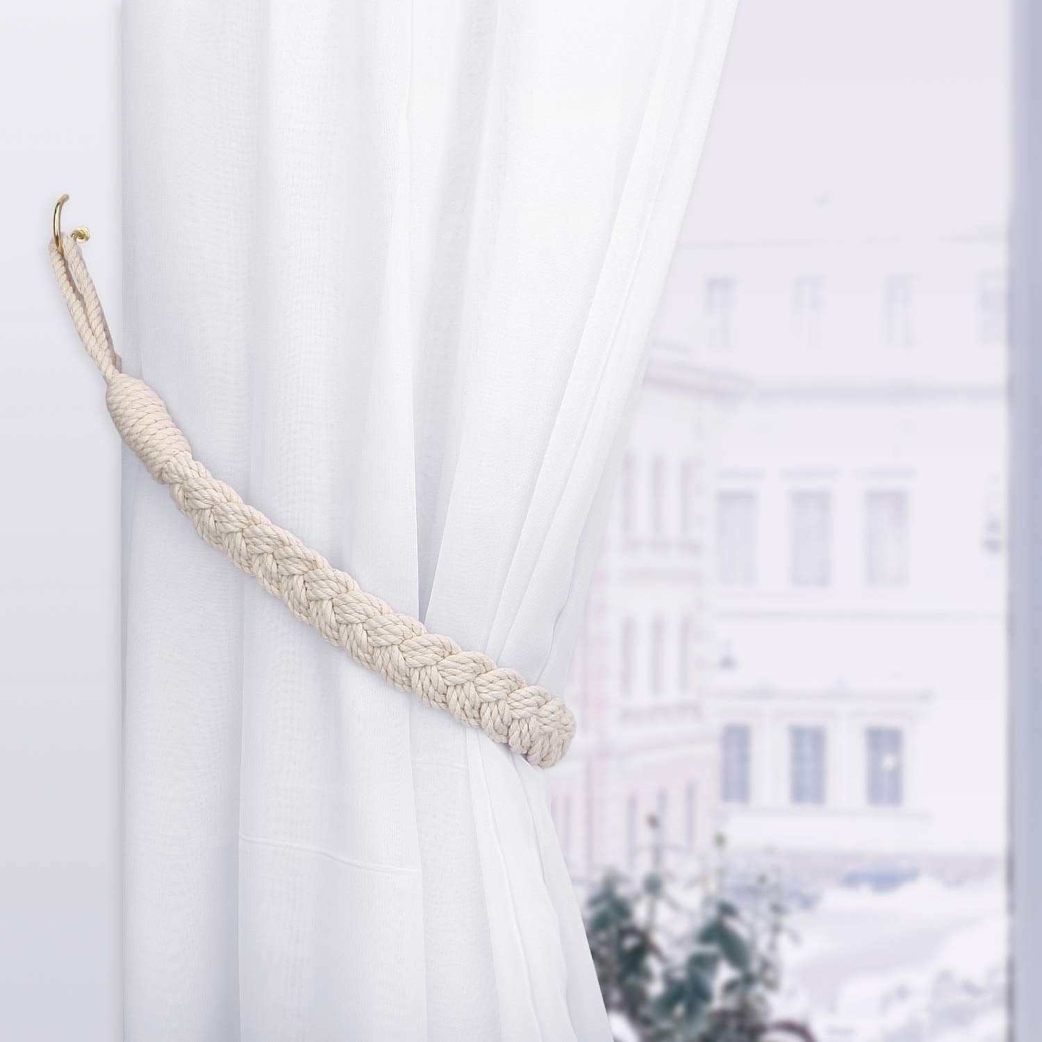 Cream braided cotton rope curtain tieback holding back a sheer white curtain, fixed to a gold wall hook for vintage style decoration.