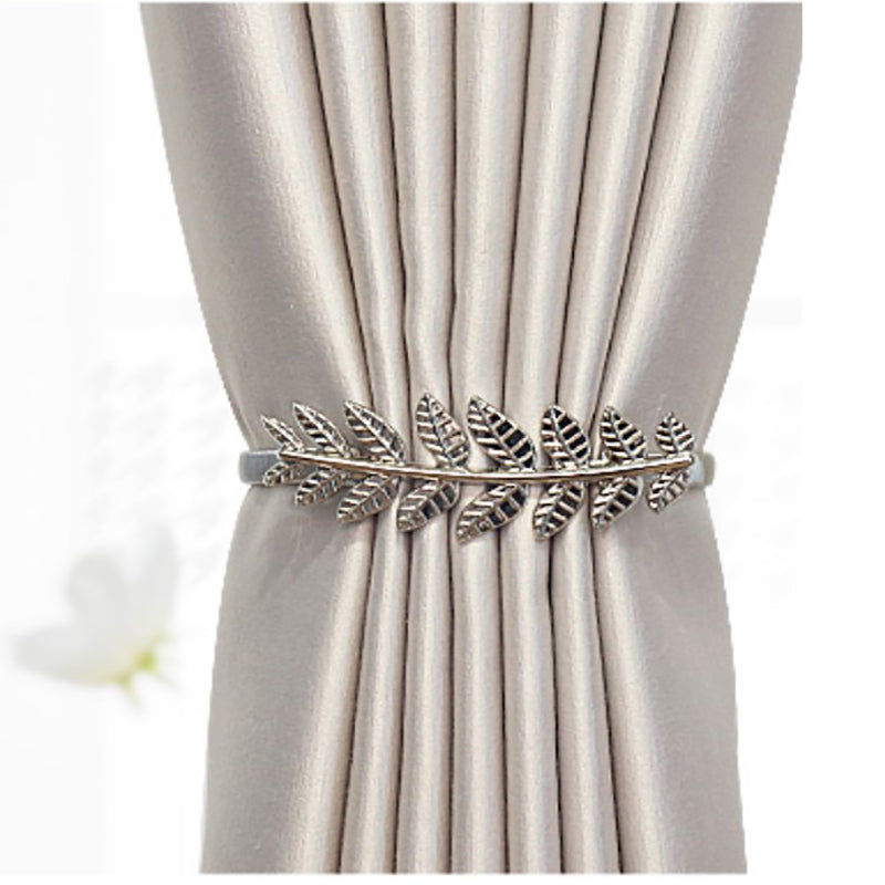 Close-up of a decorative silver metal leaf design curtain tieback holding back a beige pleated curtain panel. Elegant curtain strap.