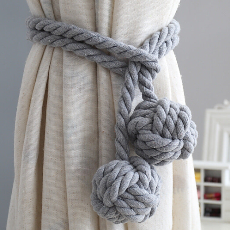 Grey twisted rope macrame curtain tieback with two decorative monkey's fist knots, securing a light-colored outdoor curtain.