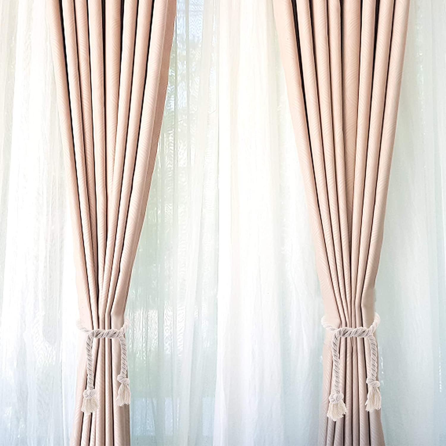 Two pleated beige outdoor curtains cinched with white braided macrame curtain tiebacks featuring decorative tassels.