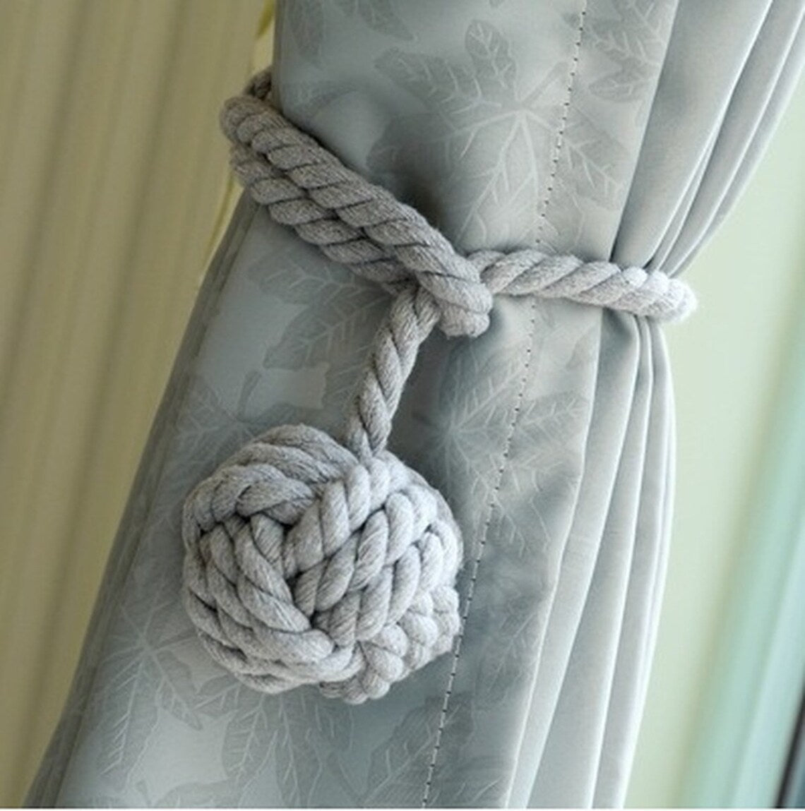 Light gray macrame rope curtain tieback with a decorative knot holding back a light blue patterned outdoor curtain.