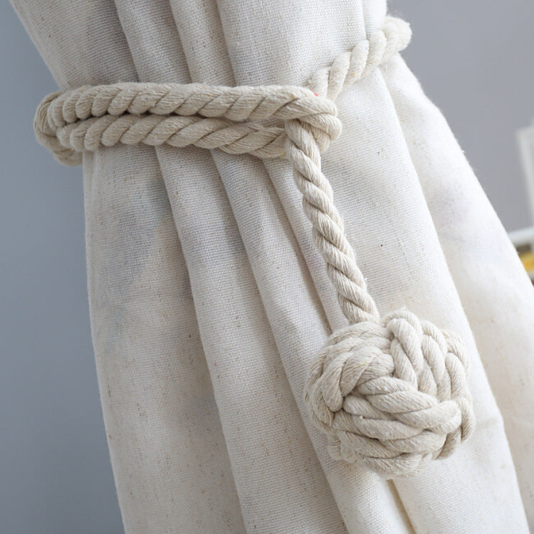 Braided natural rope macrame curtain tieback with a large decorative knot, securing a textured cream outdoor curtain.