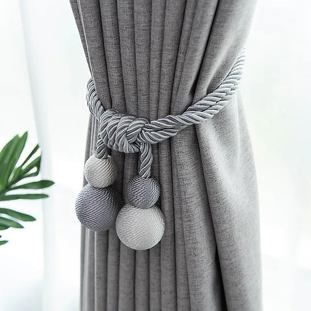 Decorative grey magnetic ball curtain tie back featuring braided rope and two-tone spheres, securing a pleated grey window curtain.