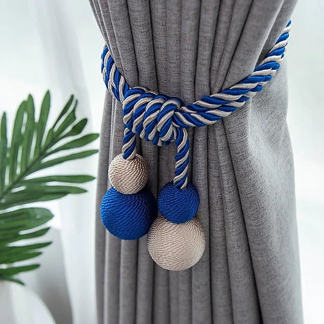 Blue & white braided magnetic curtain tieback with blue & beige decorative ball tassels cinching a grey window treatment.