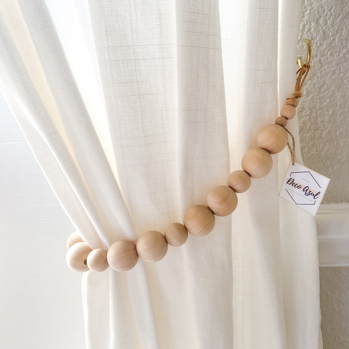 Varying natural wooden beads form a stylish curtain tieback, elegantly gathering a white textured drape for indoor or outdoor use.