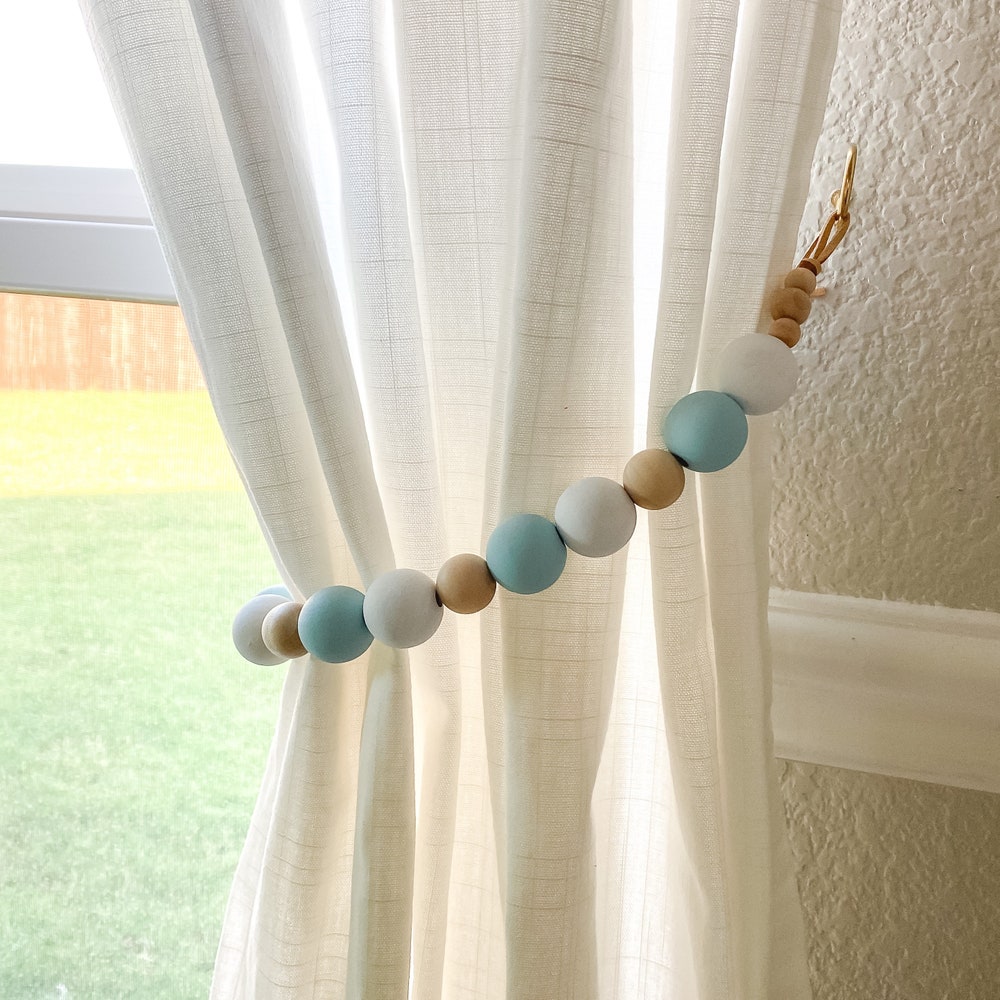Decorative wooden bead curtain tieback securing a white curtain. Features blue, white, natural wood beads & brass hook.