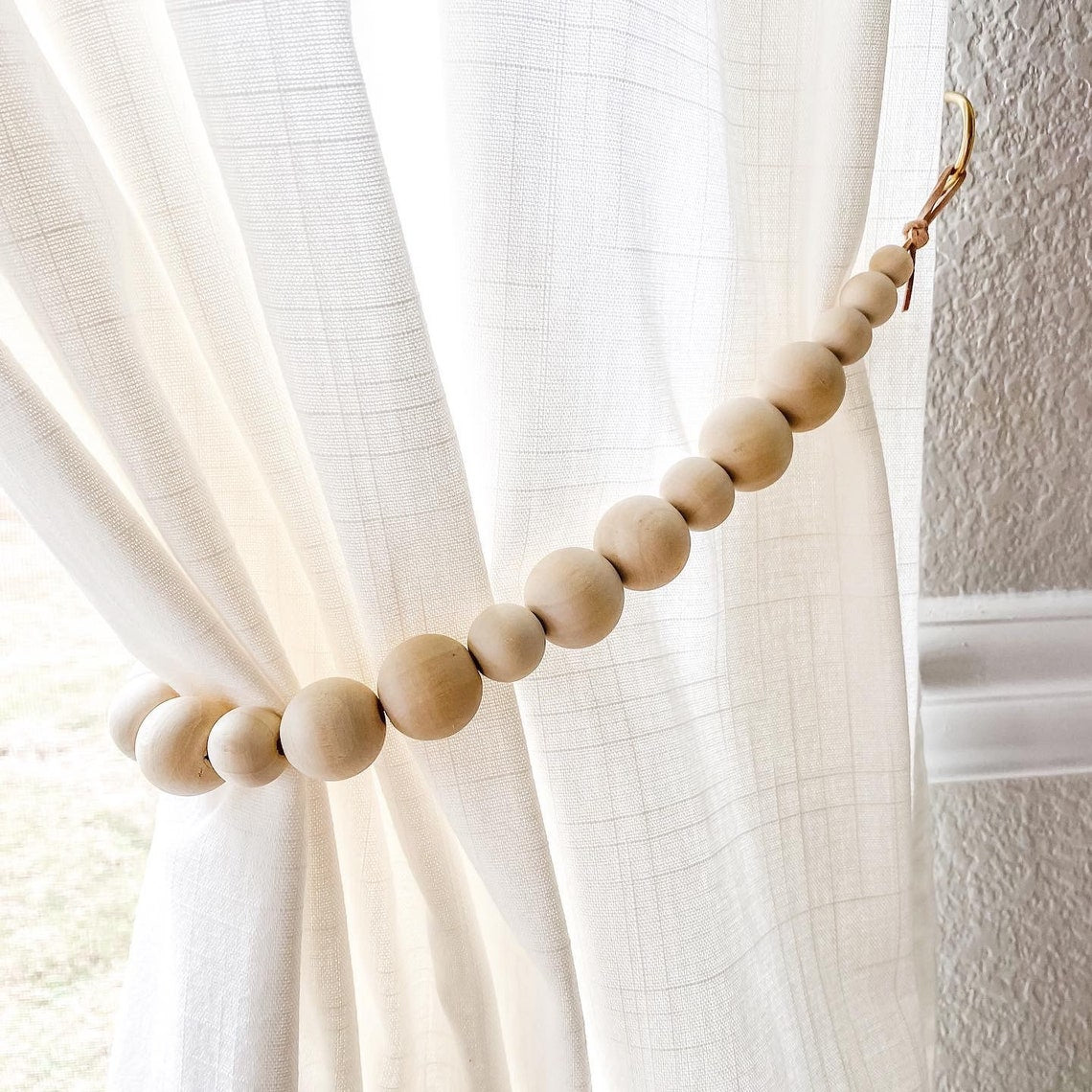 Stylish wooden bead curtain strap tieback elegantly holding a white textured curtain, secured to a wall hook.