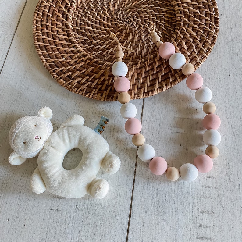 Decorative wooden beads curtain tieback with alternating pink, white, and natural wood beads, on a light wooden surface with woven rattan.