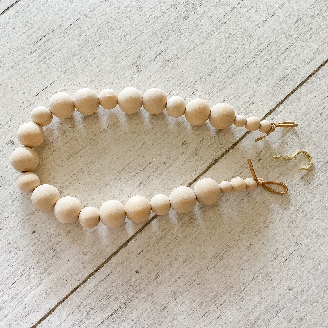 Natural wooden bead curtain strap tieback featuring varied bead sizes, leather loops, and a brass hook. Ideal for outdoor patio curtains.