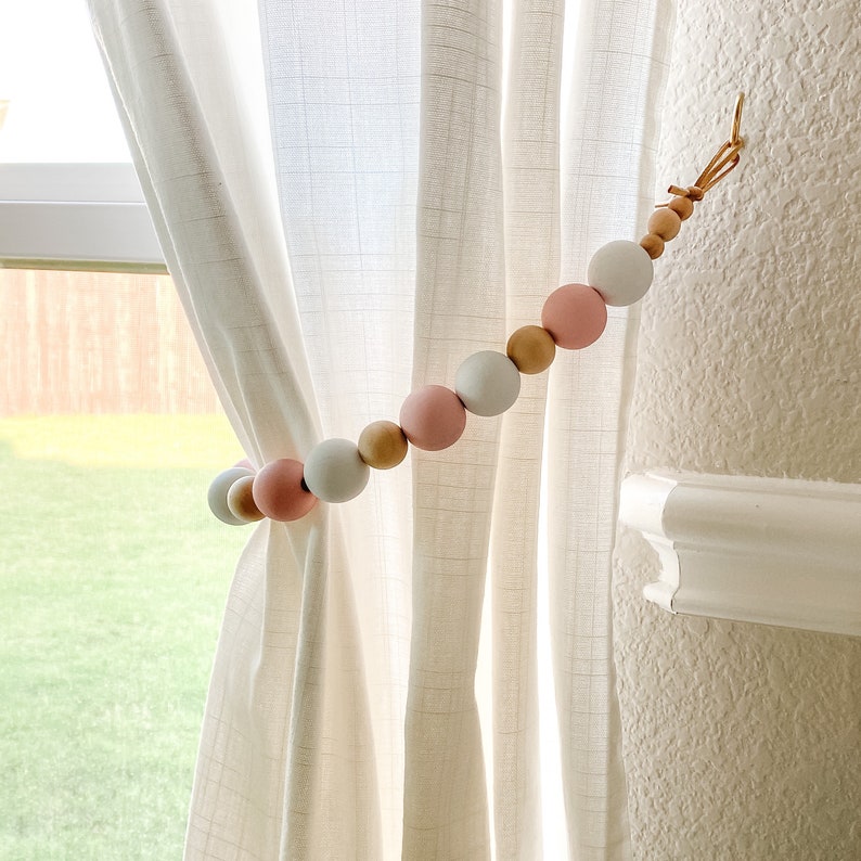 Decorative wooden beads curtain tieback with pink, white, and natural wood tones, holding a sheer curtain open.