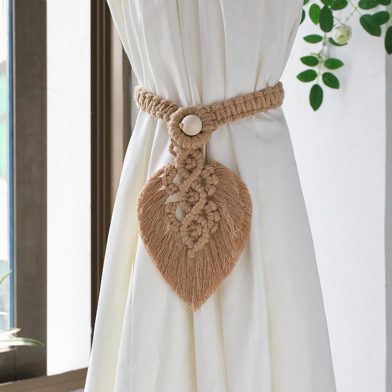 Stylish beige macrame leaf curtain tieback securing a white outdoor patio curtain, featuring braided rope and wooden bead.