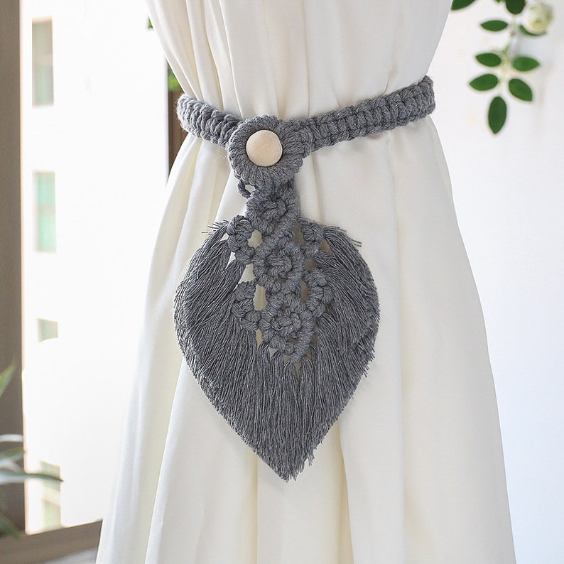 Grey macrame leaf curtain tieback with tassels and wooden button, holding a white curtain. Enhances patio, pergola, or indoor spaces.