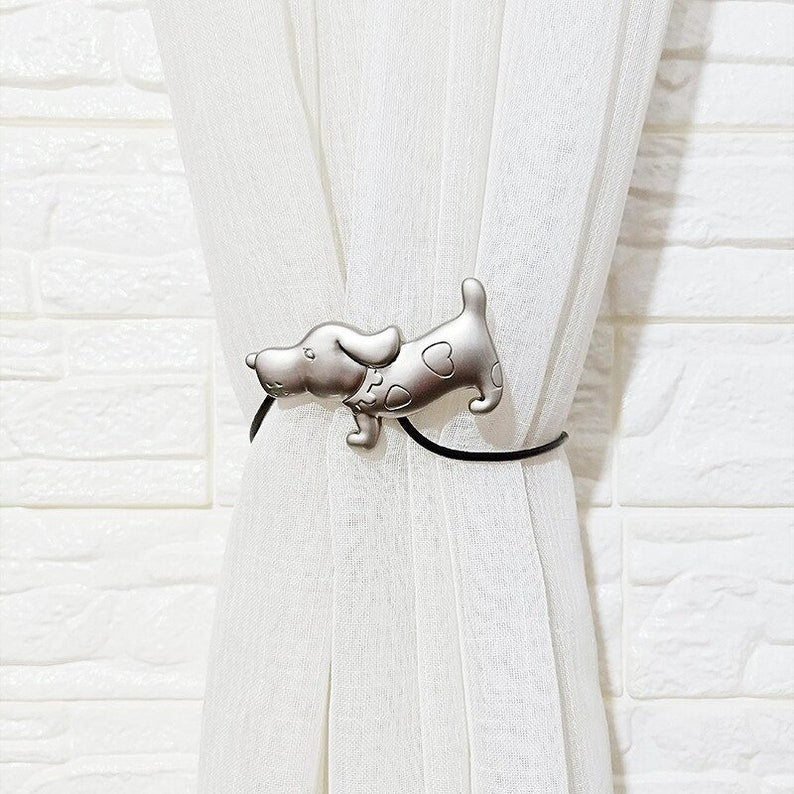 Silver dog magnetic curtain tieback with heart spots, securing a pleated white sheer curtain against a brick wall.