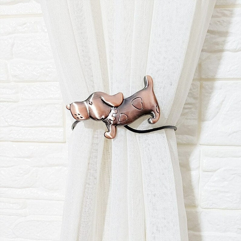 Antiqued bronze dog magnetic curtain tieback securing a sheer white curtain panel, against a textured white brick wall.