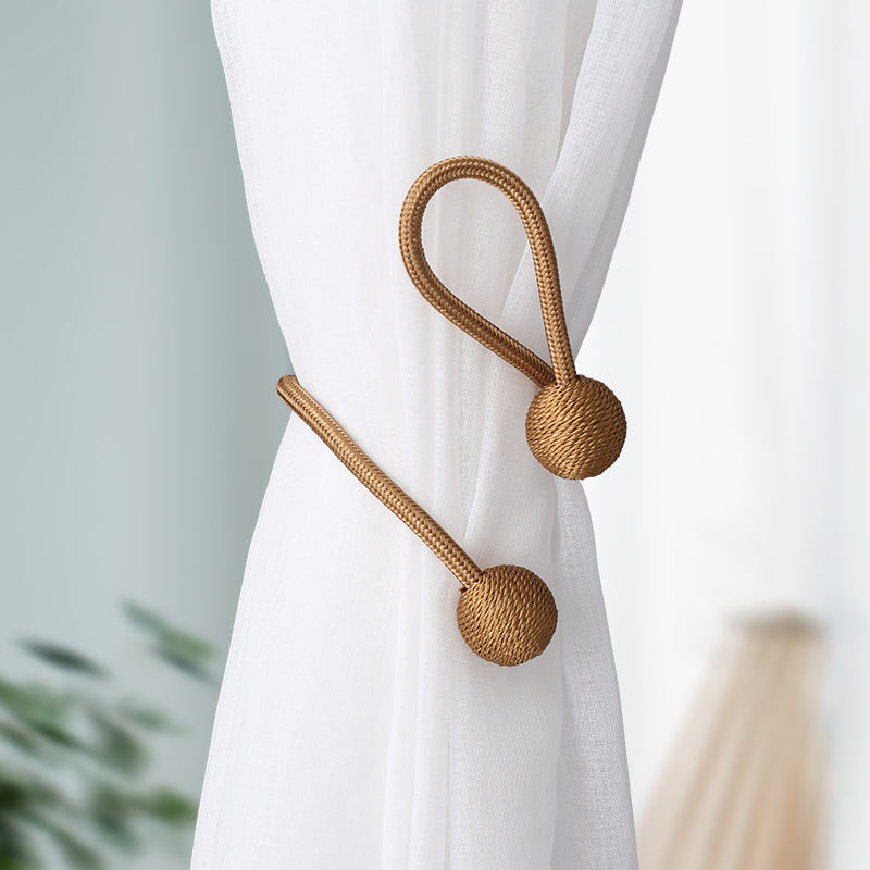 Two modern bronze magnetic curtain tiebacks, featuring braided cords and sphere ends, neatly securing a white sheer outdoor curtain.