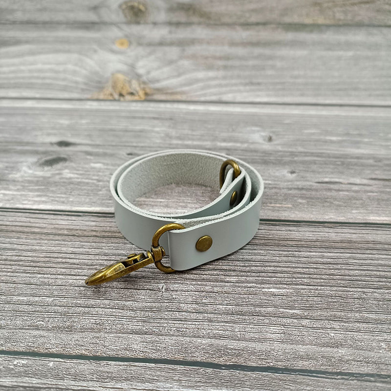Single light gray leather buckle curtain strap with antique brass tieback hardware on a weathered wooden surface.