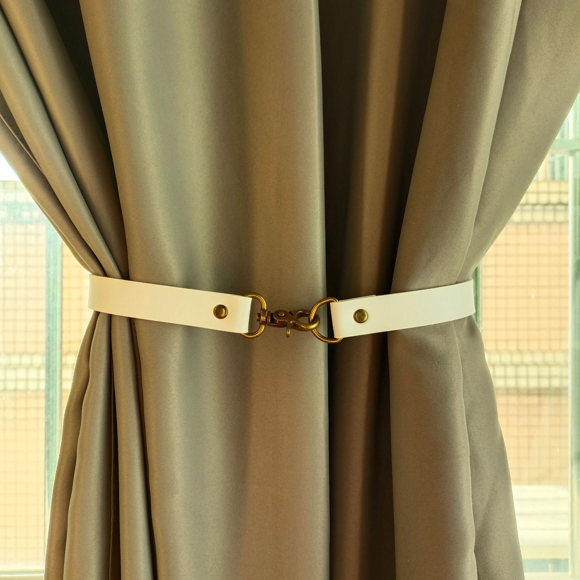 Decorative white leather buckle curtain strap tieback featuring brass hardware and studs, elegantly securing a beige outdoor curtain.