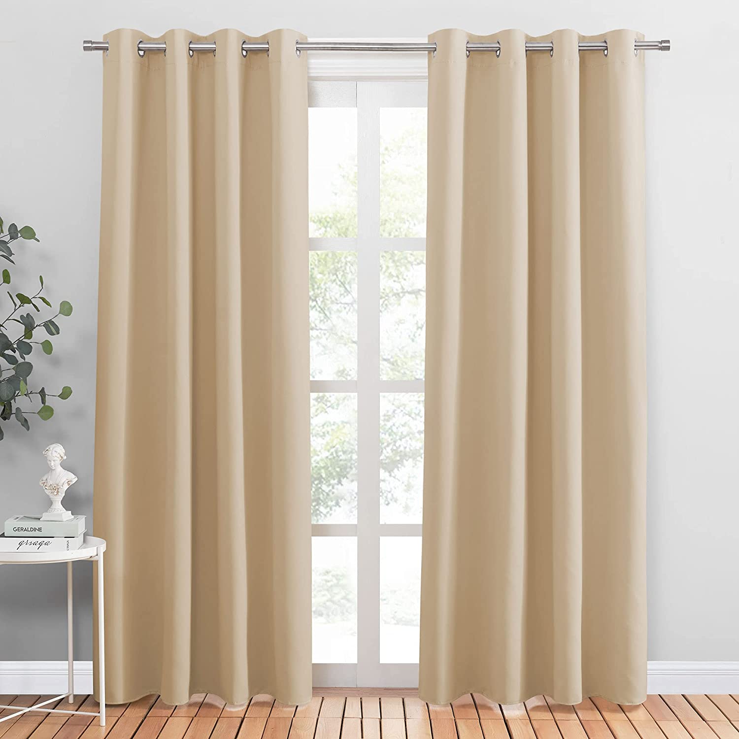 Solid beige noise reducing blackout curtains, two panels with silver grommets, hung over a window in a bright living room.