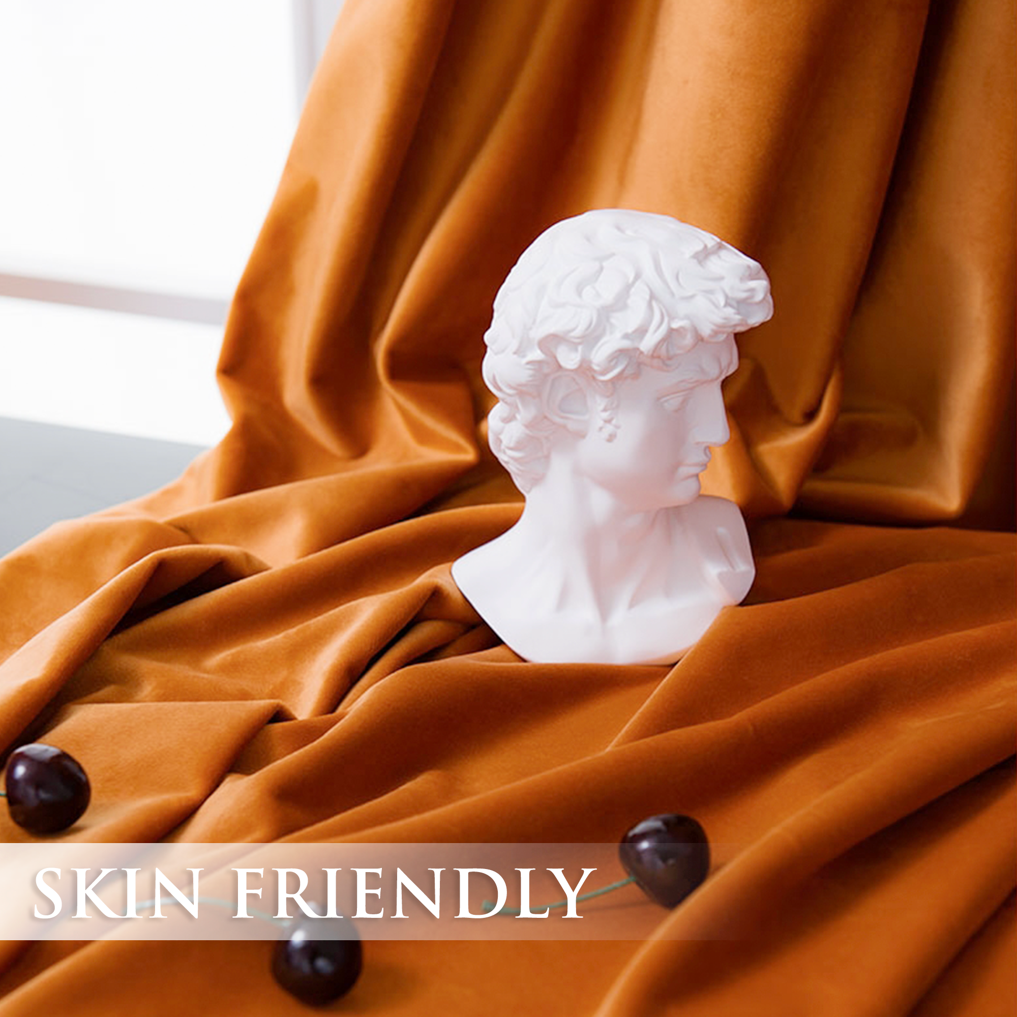 Luxurious rust orange velvet blackout curtain fabric, draped with a white bust and cherries, showcasing its skin-friendly texture.
