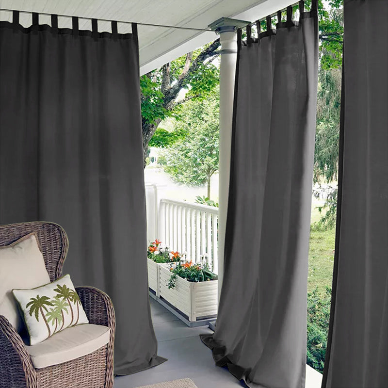 Charcoal tab top waterproof outdoor curtains creating privacy on a deck or porch. A wicker chair with a palm pillow is nearby.