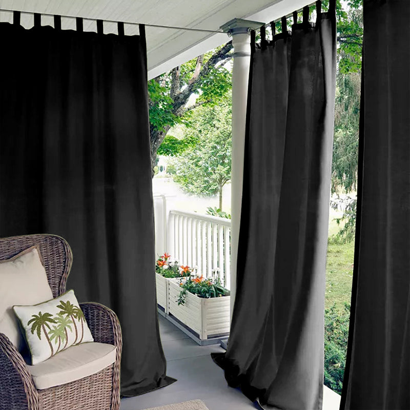 Black tab top waterproof outdoor curtains providing shade and privacy on a cozy porch deck with a wicker chair and potted flowers.