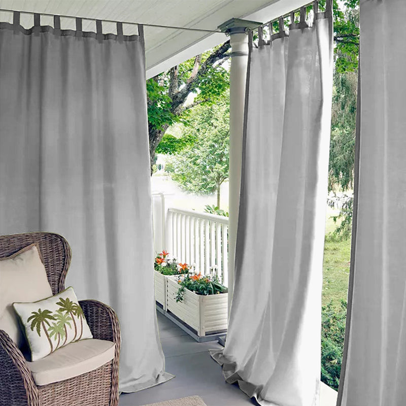 Light grey waterproof tab top outdoor curtains providing privacy on a porch deck, with a wicker chair & plants.