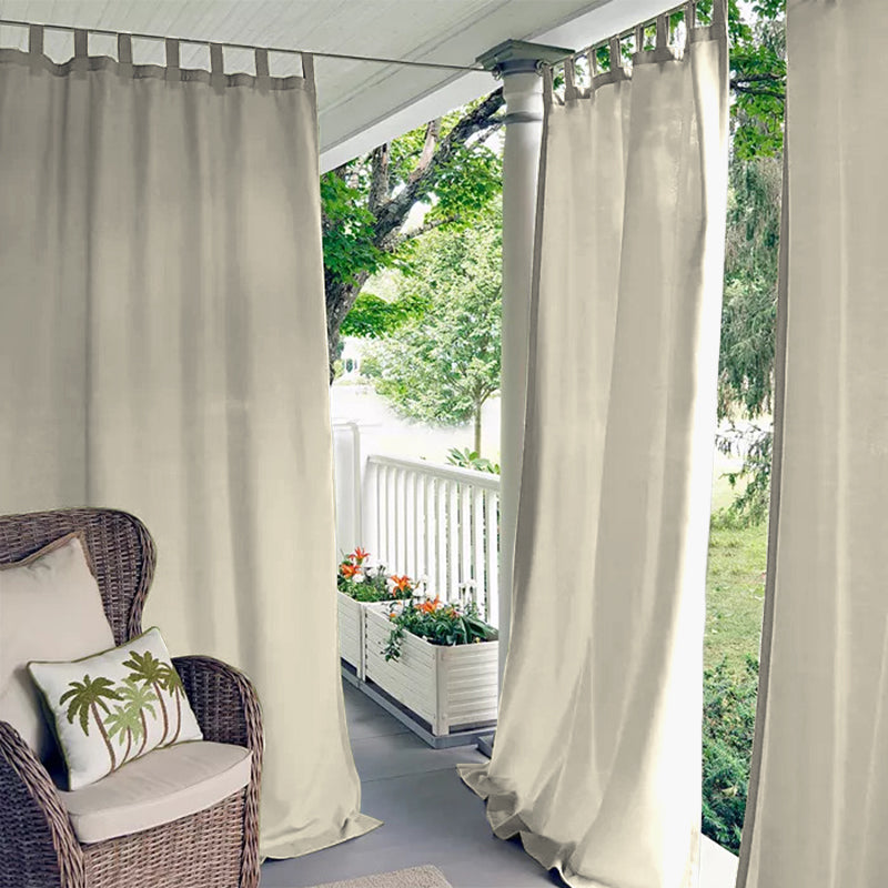 Beige waterproof tab top outdoor curtains on a porch deck, providing privacy for a wicker chair with palm pillow.
