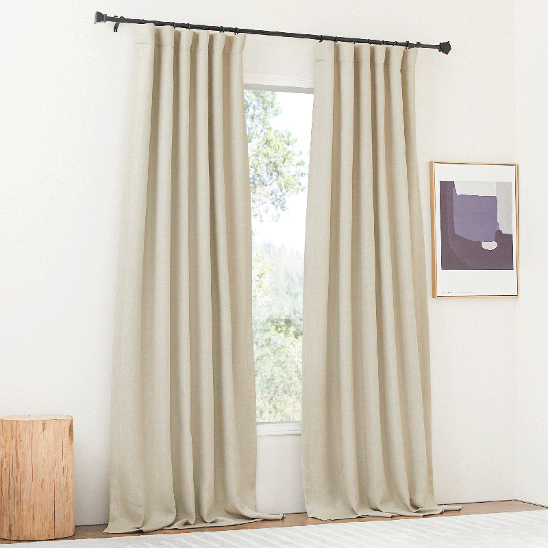Pair of plain beige rod pocket back tab blackout curtains for living room or bedroom, partially covering a window.