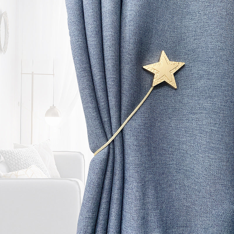 Blue textured curtain secured by a golden metal star magnetic tieback with a slender strap. Elegant magnetic curtain strap detail.