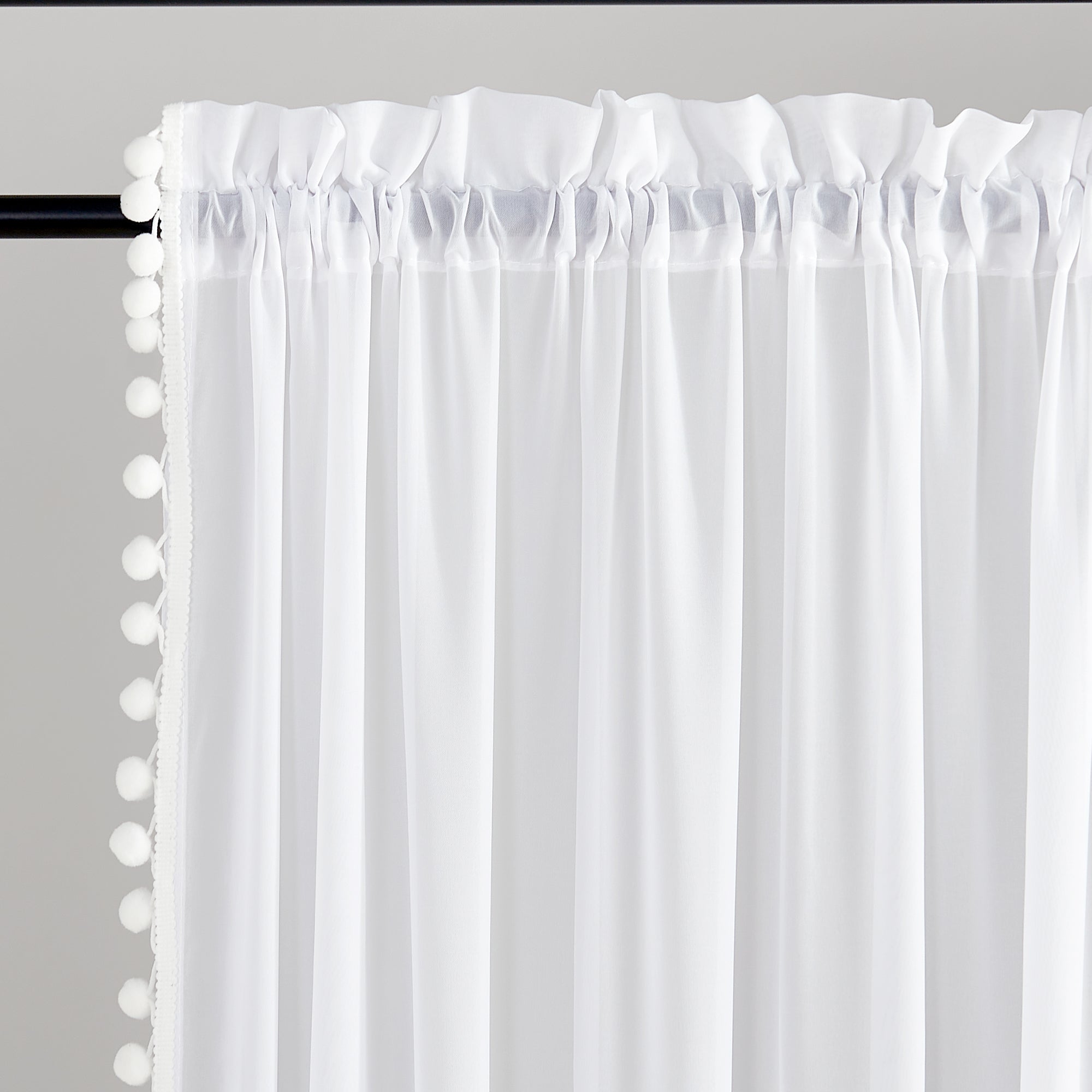 White thermal insulated blackout rod pocket curtains with playful pom pom trim, neatly gathered on a black curtain rod.