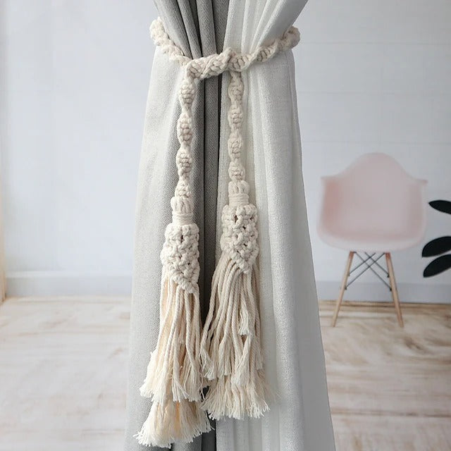 Hand-woven macrame cotton curtain tieback with two decorative tassels securing a light grey window curtain. Boho window treatment accessory.