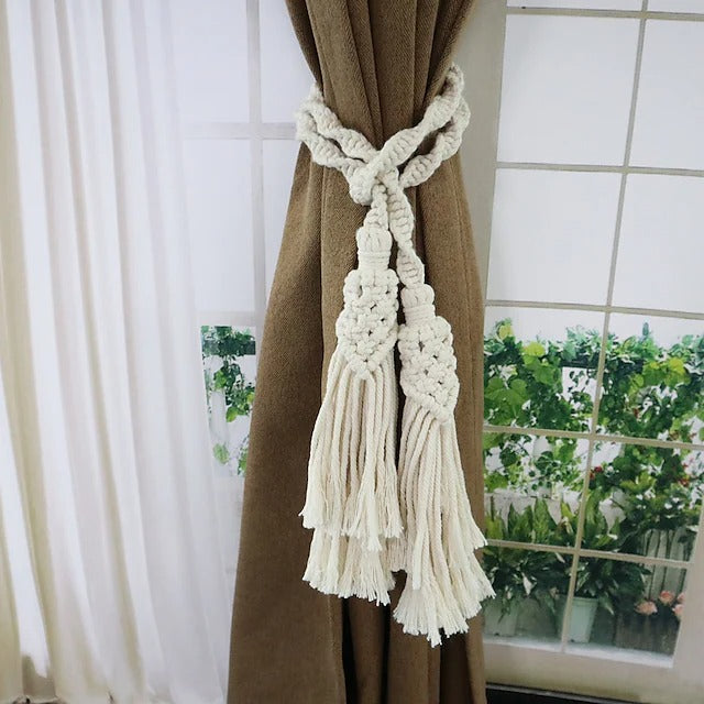 Cream hand-woven macrame curtain tieback with braided rope and double tassels, securing a brown window treatment.