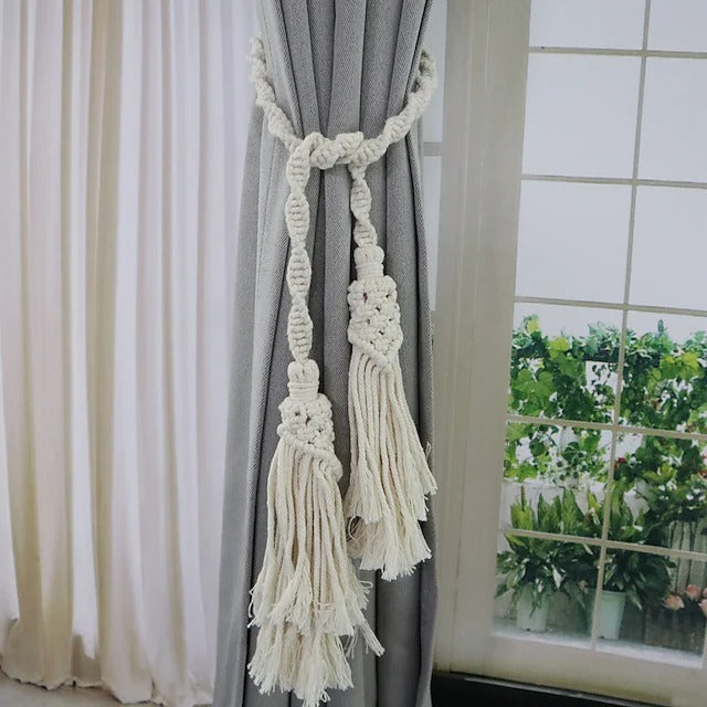 Cream hand woven macrame tassel tying rope tieback holding a grey curtain. Elegant window treatment accessory.